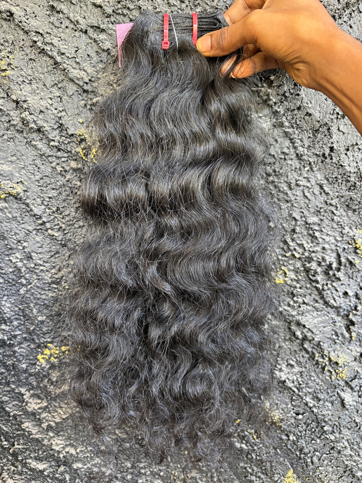 A bundle of Natural Black, Loose curly Indian human hair extensions held by a hand against a textured gray concrete wall.